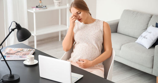 A pregnant woman at a desk looking tired and stressed, surrounded by papers, a laptop, and a cup of coffee. Her posture shows fatigue, symbolizing stress during pregnancy in an office setting.