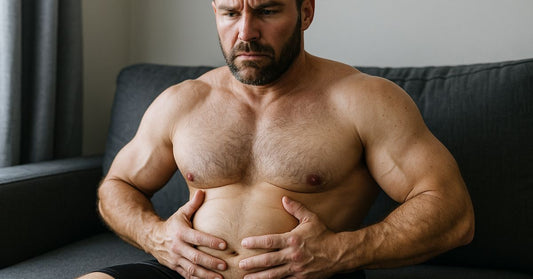 A muscular man, shirtless, sitting on a couch, holding his bloated stomach with a slight expression of discomfort. The image illustrates the phenomenon of water retention caused by testosterone, set in a relaxed setting